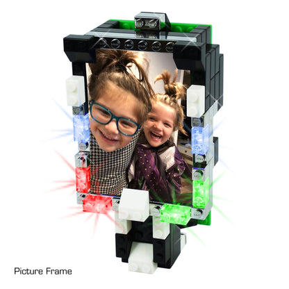 A rectangular picture frame constructed with E-Blox's Power Blox™ Builder 292 Classroom Set - LED Light-Up Building Blocks showcases a photo of two smiling children, one of whom is wearing glasses. The frame, in line with STEM principles, features illuminated sections in red, green, and blue. The base comprises additional plastic bricks and carries the label "Picture Frame" below.