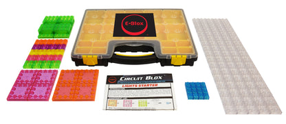 Image shows a yellow and black carrying case labeled "E-Blox," surrounded by various translucent building blocks in green, red, pink, yellow, blue, and clear. A white instruction sheet titled "Circuit Blox™ Sound Activated Dancing Lights Classroom Set - E-Blox®" is included in this educational play STEM kit featuring colorful lights.