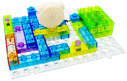 The Circuit Blox™ 120 Project Classroom Set by E-Blox features a multicolored, translucent building block set on a clear base, with components in blue, green, yellow, and purple. It includes plastic blocks, a white gear, and an attached speaker or motor. This set is perfect for STEM programs and meets NGSS requirements.