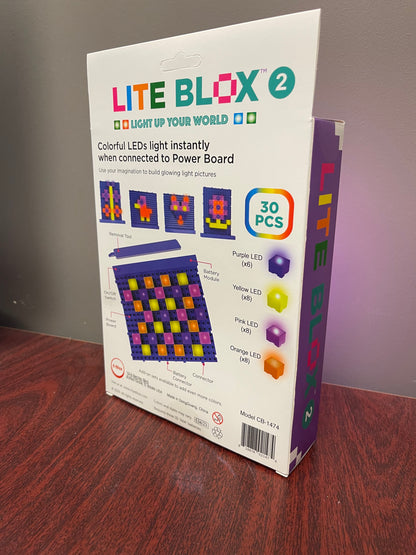 A box labeled "Lite Blox 2 - Light up your world with all new colors!" from E-Blox, Inc. sits on a wooden surface against a gray background. The packaging showcases colorful LED Blox that light up when connected to a power board, featuring examples of creative designs. Inside, you'll find 30 vibrant pieces in purple, yellow, pink, and orange.