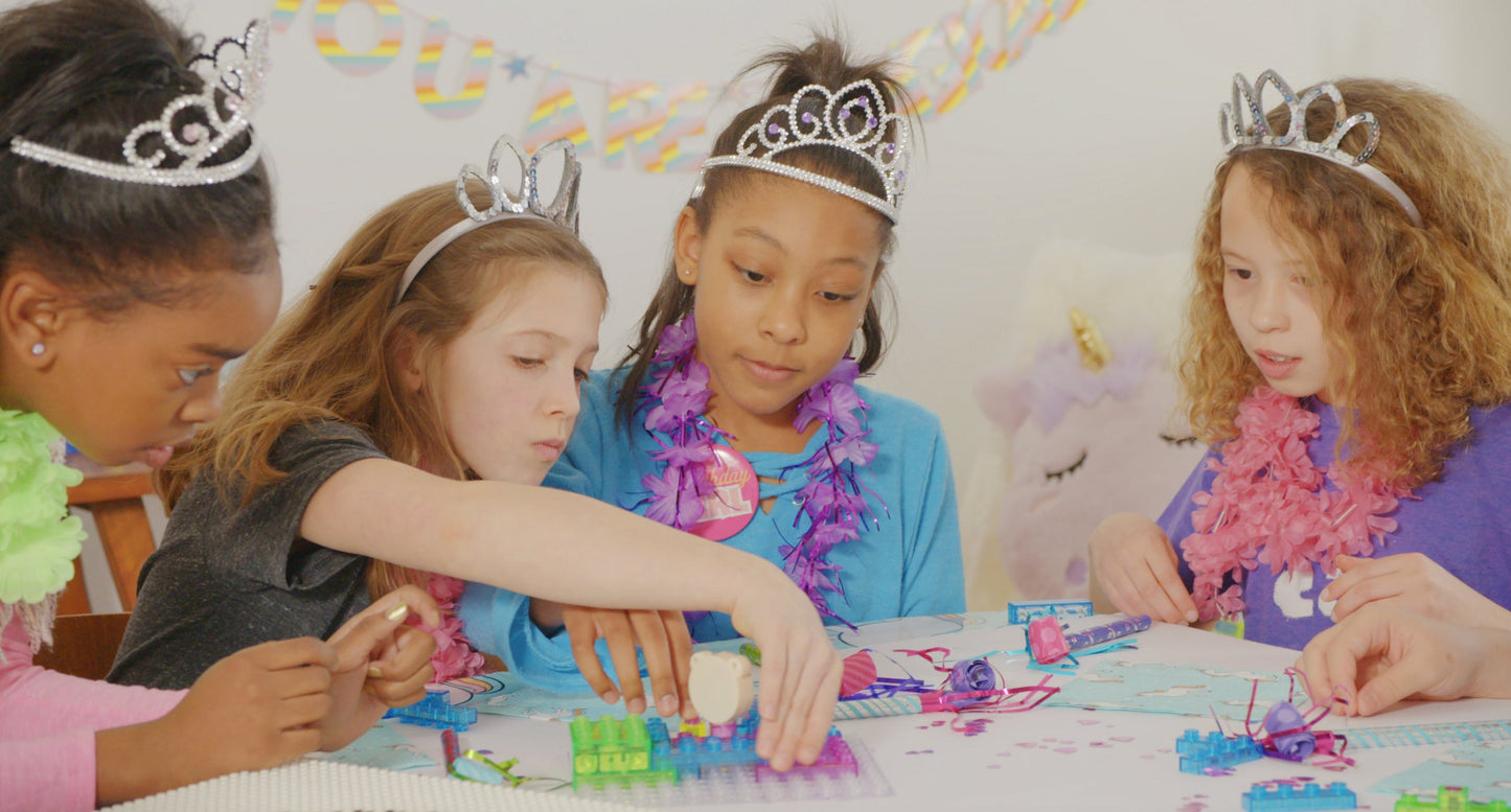 Four girls wearing tiaras and colorful feather boas sit around a table, deeply engrossed in constructing projects with the Circuit Blox™ 59 project Classroom Set - E-Blox® Circuit Board Building Blocks Educational Sets. Their focused and engaged expressions embody the spirit of STEM education, with a banner in the background that says "You are special.