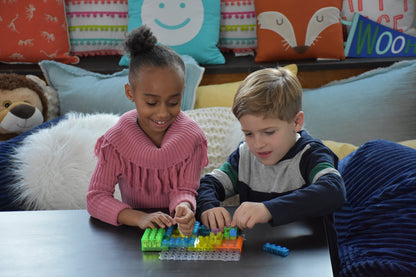 Two children are playing with the Circuit Blox™ 59 project Classroom Set by E-Blox on a black table. The girl, in a pink sweater, and the boy, in a blue-striped shirt, are engrossed in assembling the circuit board building blocks. Surrounded by cushions and plush toys, their playtime subtly incorporates principles of STEM education.