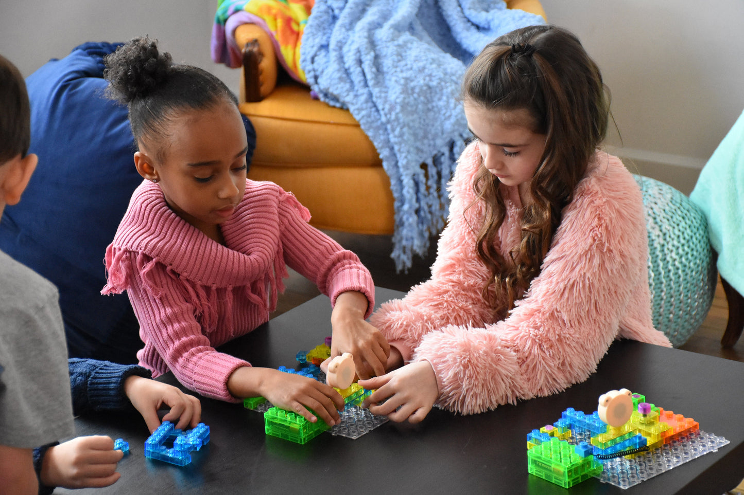 Two young girls sit at a table, focused on building with the colorful Circuit Blox™ 120 Project Classroom Set from E-Blox. One girl wears a pink knit sweater, and the other wears a fluffy pink jacket. A blue blanket and a brown chair are in the background. Their project aligns perfectly with NGSS requirements for an engaging STEM program.