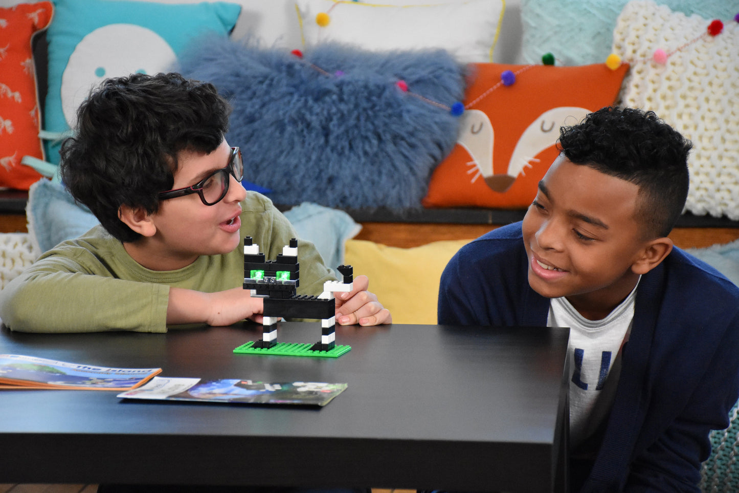 Two boys sit at a table playing with a black and white toy, likely the Power Blox™ Builds Plus Set - E-Blox® - LED Light-Up Building Blocks Student Set. They are smiling and looking at each other. Behind them, colorful pillows with animal designs adorn the couch. A magazine lies open on the table in front of them, possibly discussing NGSS requirements that guide their STEM curriculum activities.