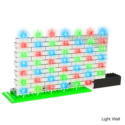 A wall constructed of interlocking white Power Blox Flashing Frenzy - E-Blox® LED light-up building blocks, featuring blue, red, and green LEDs arranged in a precise grid pattern. The base is green with a black rectangular component at the bottom right corner labeled "Light Wall." This patented technology by E-Blox can be seamlessly integrated into any STEM curriculum to teach 3D circuits.