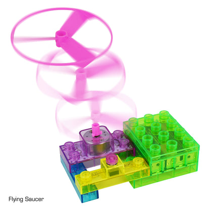 A toy flying saucer launcher with colorful translucent blocks and a pink propeller shown in motion. The bottom blocks are blue and green, featuring electronic circuits that connect to the small motor attached to the pink spinning propeller. "Flying Saucer" is labeled in the bottom left corner as part of E-Blox's Circuit Blox™ BYO Flying Saucer Circuit Board Building Blocks Toys series.