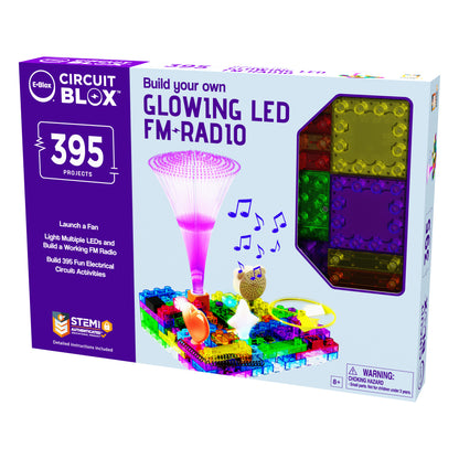 Here's an image of the Circuit Blox™ 395 - E-Blox® Circuit Board Building Blocks Toys for Kids. The predominantly white box sports a purple and green color scheme. The packaging displays "Build your own Glowing LED FM Radio" along with information about 395 building projects. The front highlights features like a glowing fan, LEDs, and musical notes to emphasize its STEM program benefits. It includes a warning indicating that it is suitable for ages 8 and up.