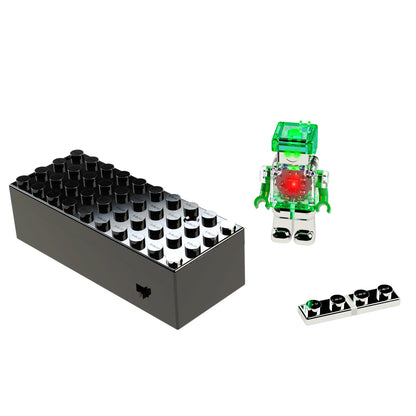 A black, rectangular LEGO brick with multiple connector knobs, a small green and transparent LUMEN®- Robyn- Illuminated Mini Figure robot by E-Blox, Inc., featuring a red circular component, and a small black LEGO piece with a row of connector knobs are displayed against a white background.