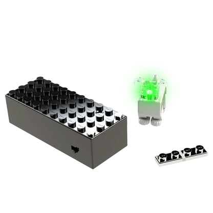 A black E-Blox® brick with a switch on its side and another small detached chrome E-Blox® brick in the foreground. A glowing green LUMEN®-Glen-Illuminated Mini Figure is hovering above the main brick. The setup, part of the Story Blox™ series by E-Blox®, appears to be electronic or battery-powered with a light feature.