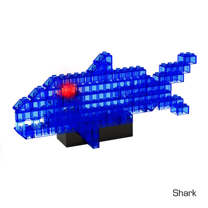 A construction made of blue transparent building blocks in the shape of a shark, with a black base and a small red block inside the structure. The word "Shark" is written in the bottom right corner. This 3D light-up structure is part of our Power Blox™ Builder 4-in-1 - E-Blox® LED Light-Up Building Blocks from E-Blox.