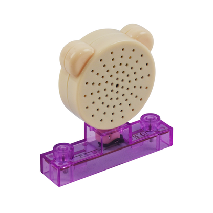 A beige, bear-shaped speaker with small perforations on the front, resting on a transparent purple plastic stand. The stand features two protruding "studs" typical of interlocking building blocks and is part of the E-Blox Speaker Block for Circuit Builder.