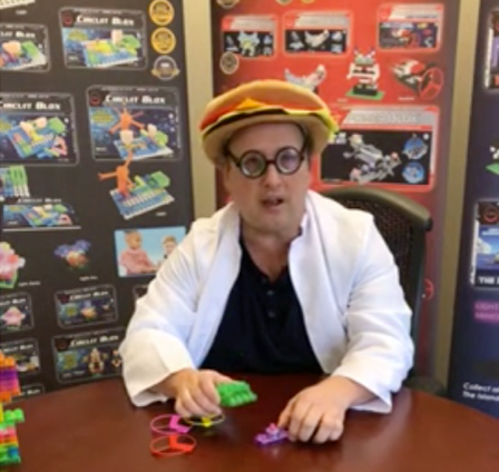 A person wearing a white lab coat, round glasses, and a hat resembling a hamburger sits at a table with E-Blox e-Learning Session 1 sets by E-Blox, Inc., surrounded by colorful building blocks and educational materials. Informational boards behind them display various construction sets.