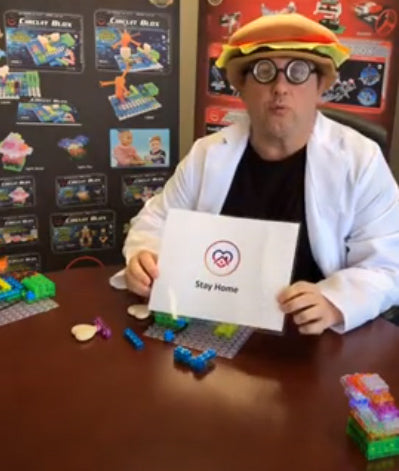 A person in a white lab coat and a hamburger hat with round glasses is sitting at a table covered with colorful E-Blox building blocks. They are holding a sign that reads "Stay Home" with a heart icon. The background shows posters of various E-Blox e-Learning Session 6 sets from E-Blox, Inc.