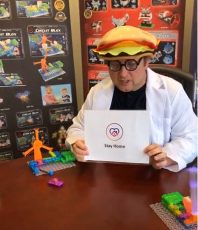 A person dressed in a white lab coat and a burger hat holds a sign that says "Stay Home" while sitting at a table with colorful E-Blox e-Learning Session 9 building blocks. The background features posters of E-Blox, Inc. toy sets.